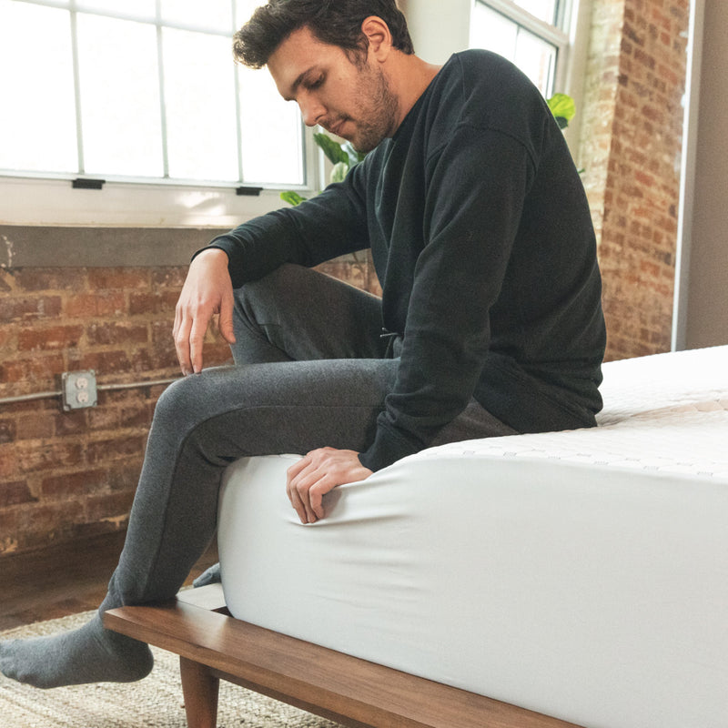 Photo of a man sitting on a corner of a bed that is fitted with a white and gray mattress protector.