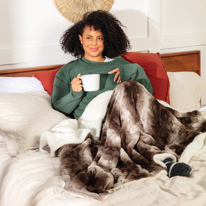 Photo of a woman smiling, holding a mug of coffee with a dark espresso brown faux fur blanket with a fluffy white sherpa on the reversed side draped over her lap.
