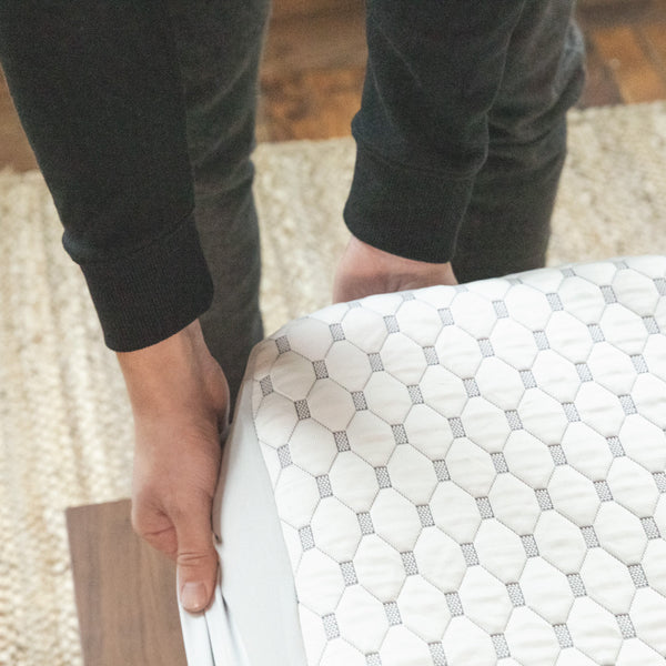 A person pulling a white and gray mattress protector over the corner of a mattress. (No Script, Alternate View)